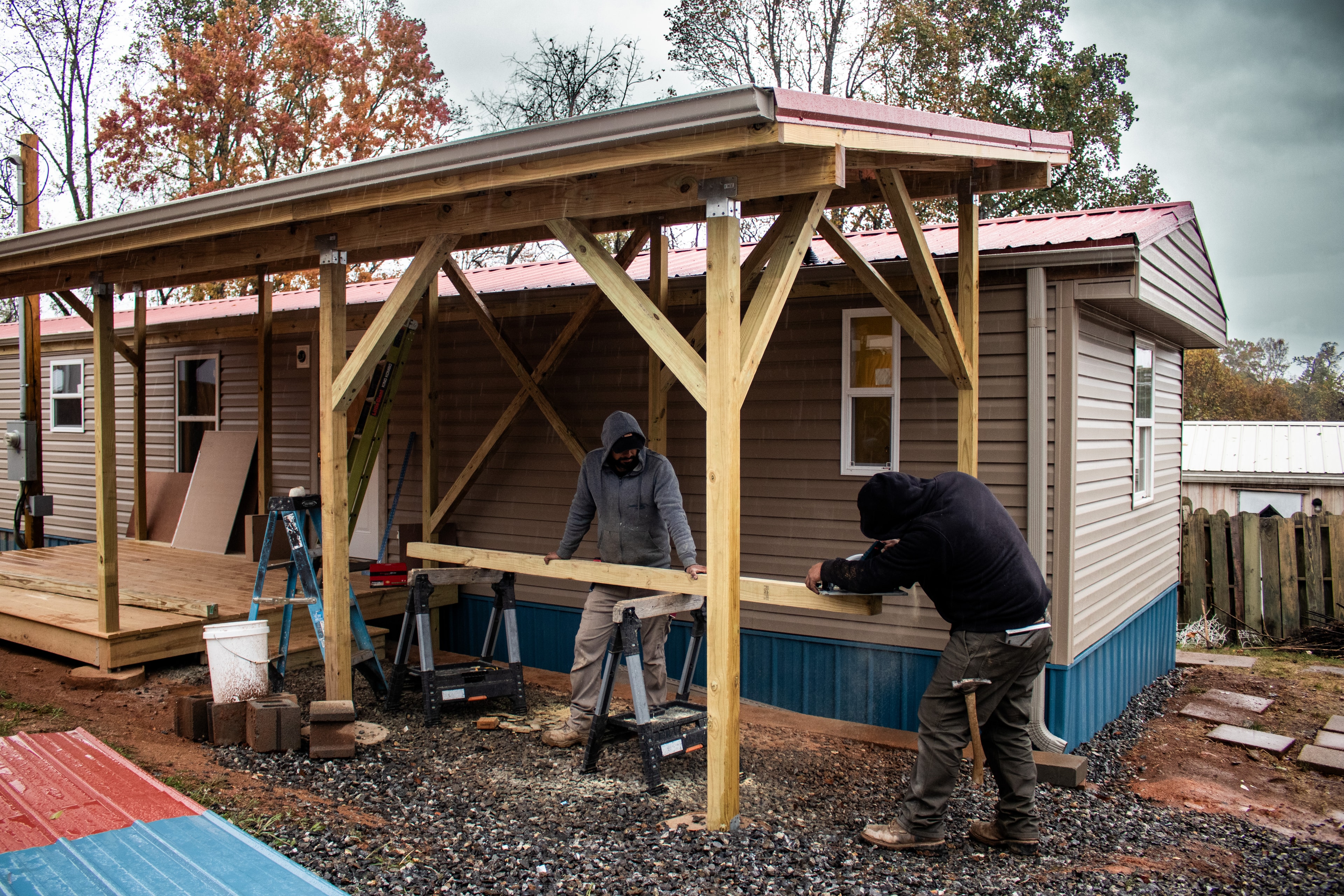 Workers cut wood under an elegant porch in front of a restored mobile home.